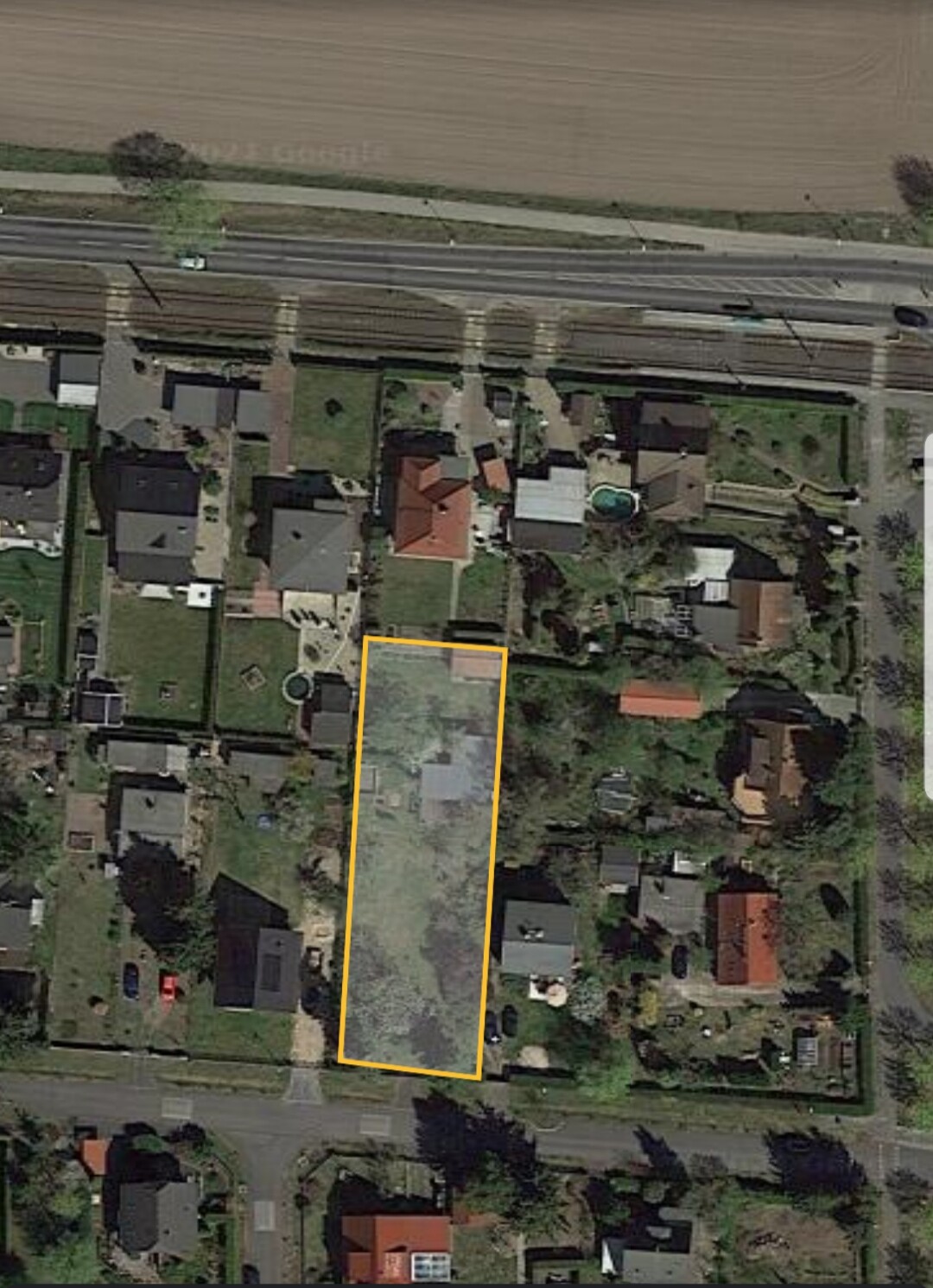Aerial view: elongated, undeveloped plot in a residential area, outlined in orange.