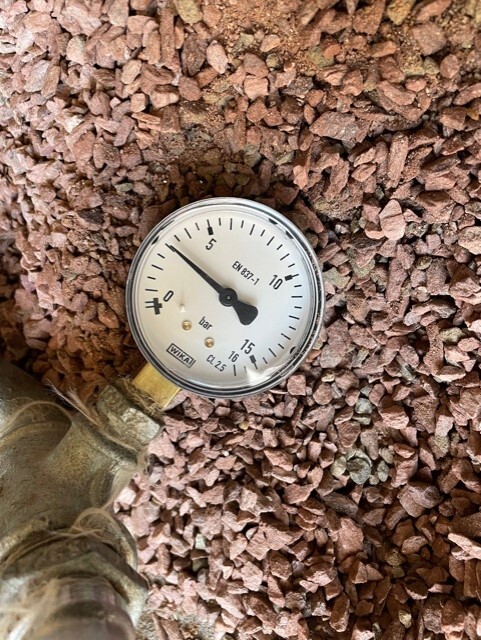 Pressure gauge with 0–16 bar scale, on a metal valve, resting on gravel.