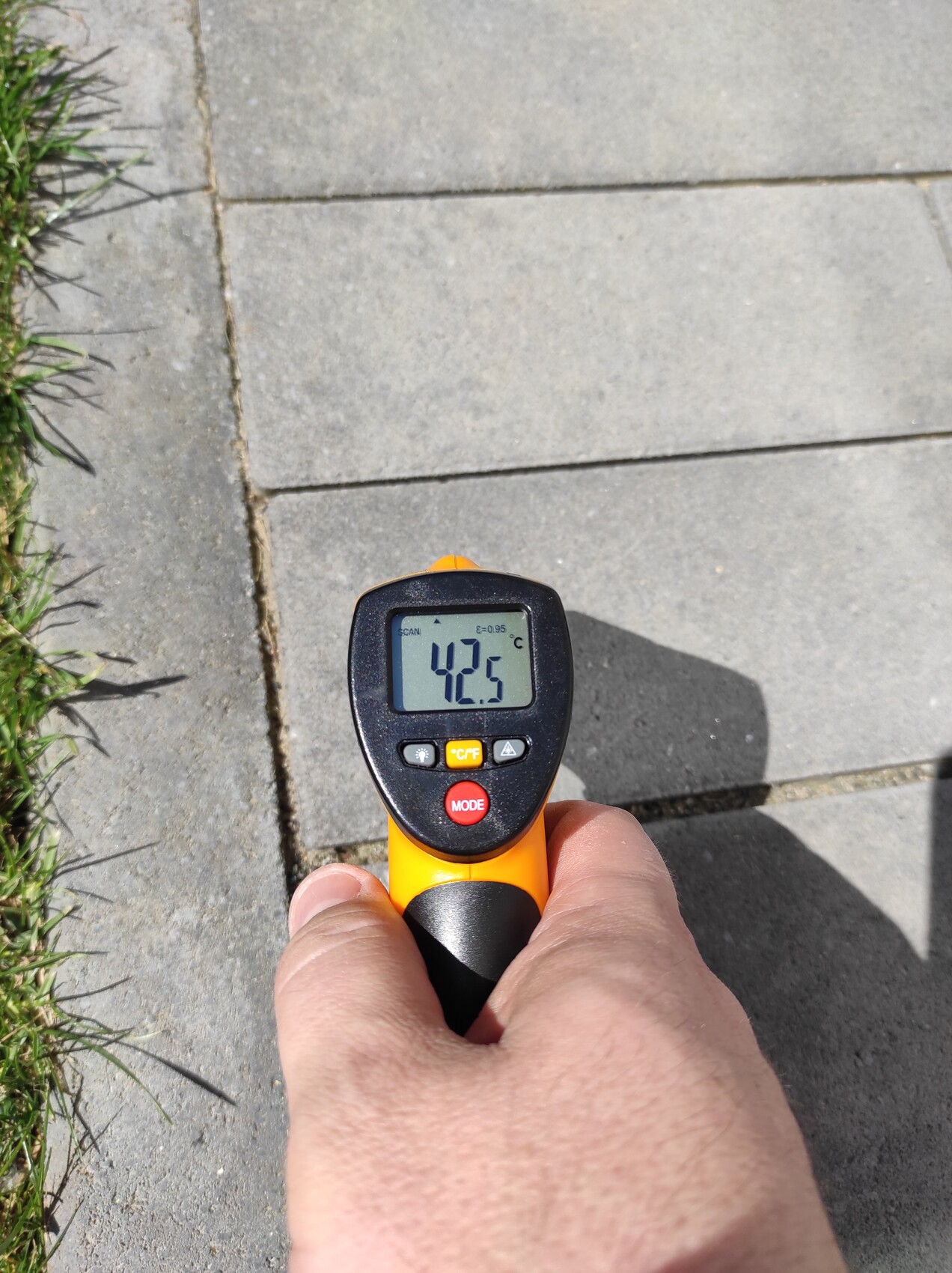 Hand holding infrared thermometer over walkway slabs; display shows 42.5°C