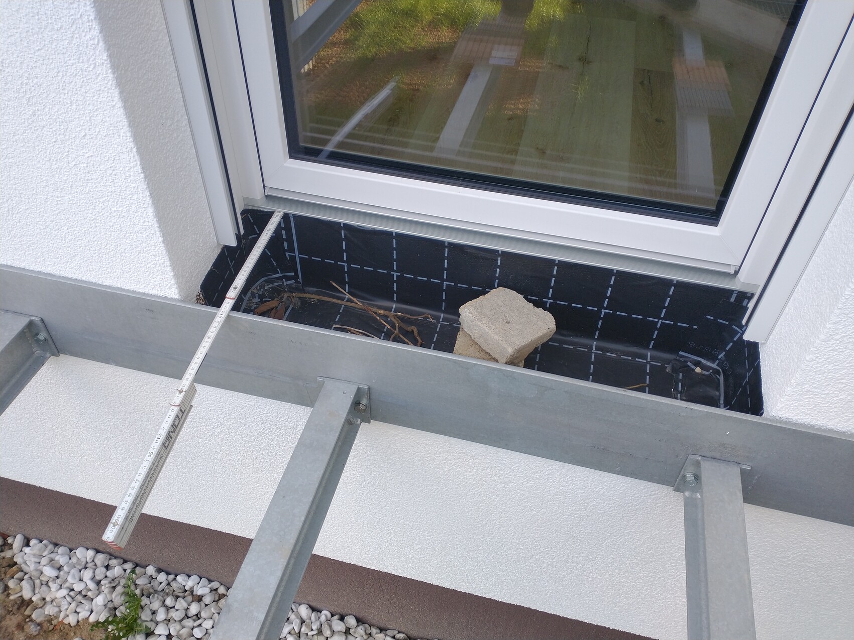 Außenfenster mit weißem Rahmen, darunter schwarze Abdichtbahn, Betonstein und Metallrahmenkonstruktion