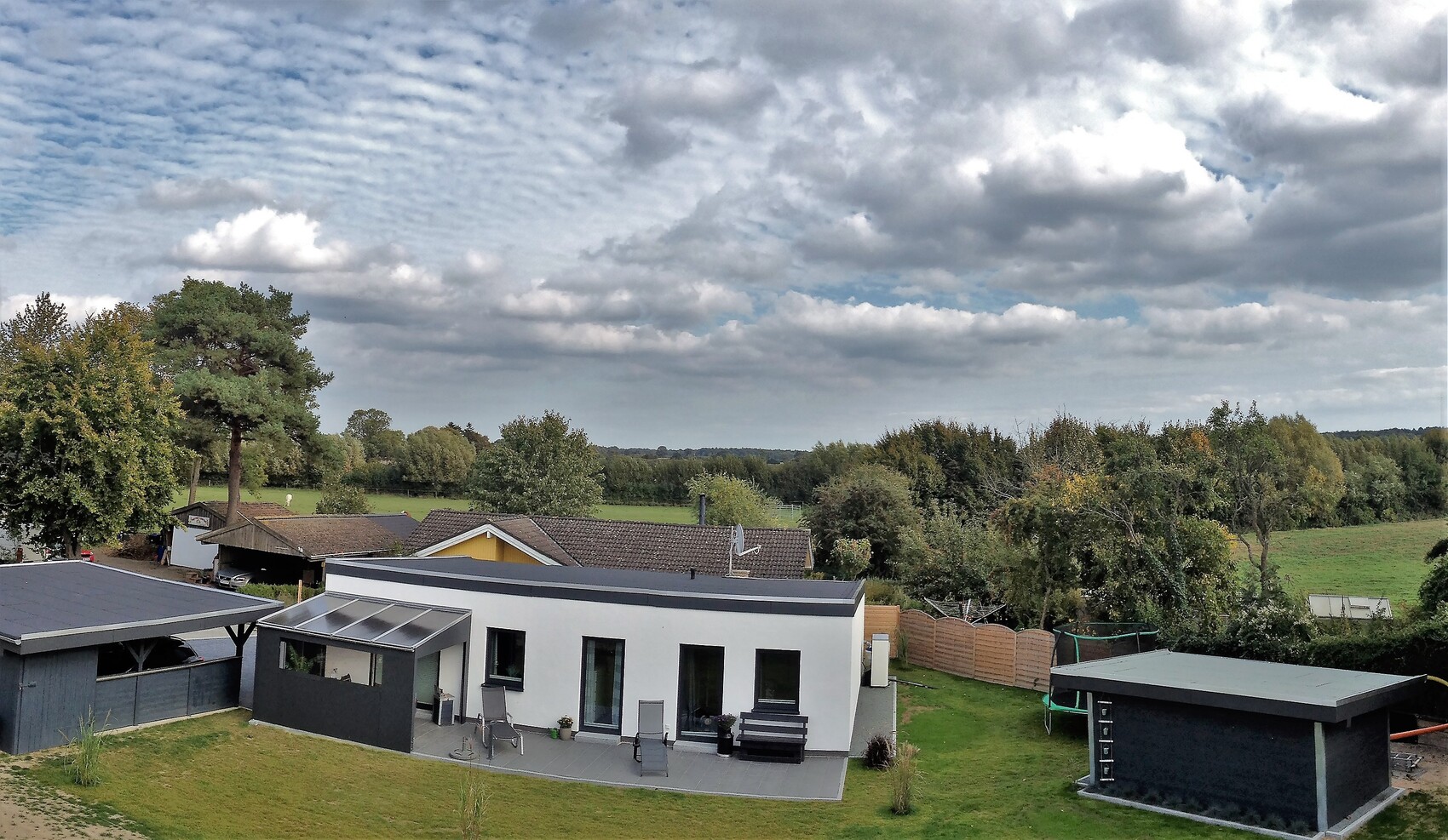 Modernes, flaches Einfamilienhaus mit Terrasse, Garten und Trampolin, Baumreihen im Hintergrund.