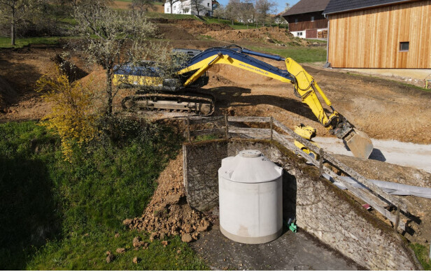 Gelber Bagger arbeitet an Baustelle am Hang, neben weißem Wassertank und Holzgeländer.
