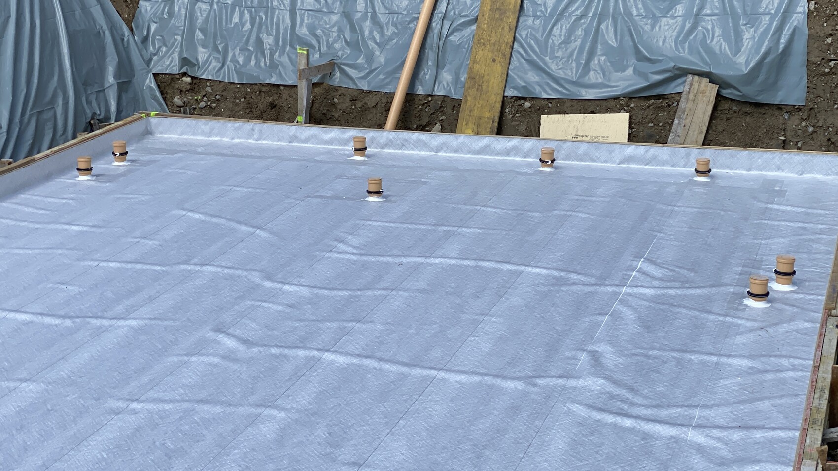 Foundation slab with blue waterproofing membrane, surrounding wooden frame, and capped pipes.
