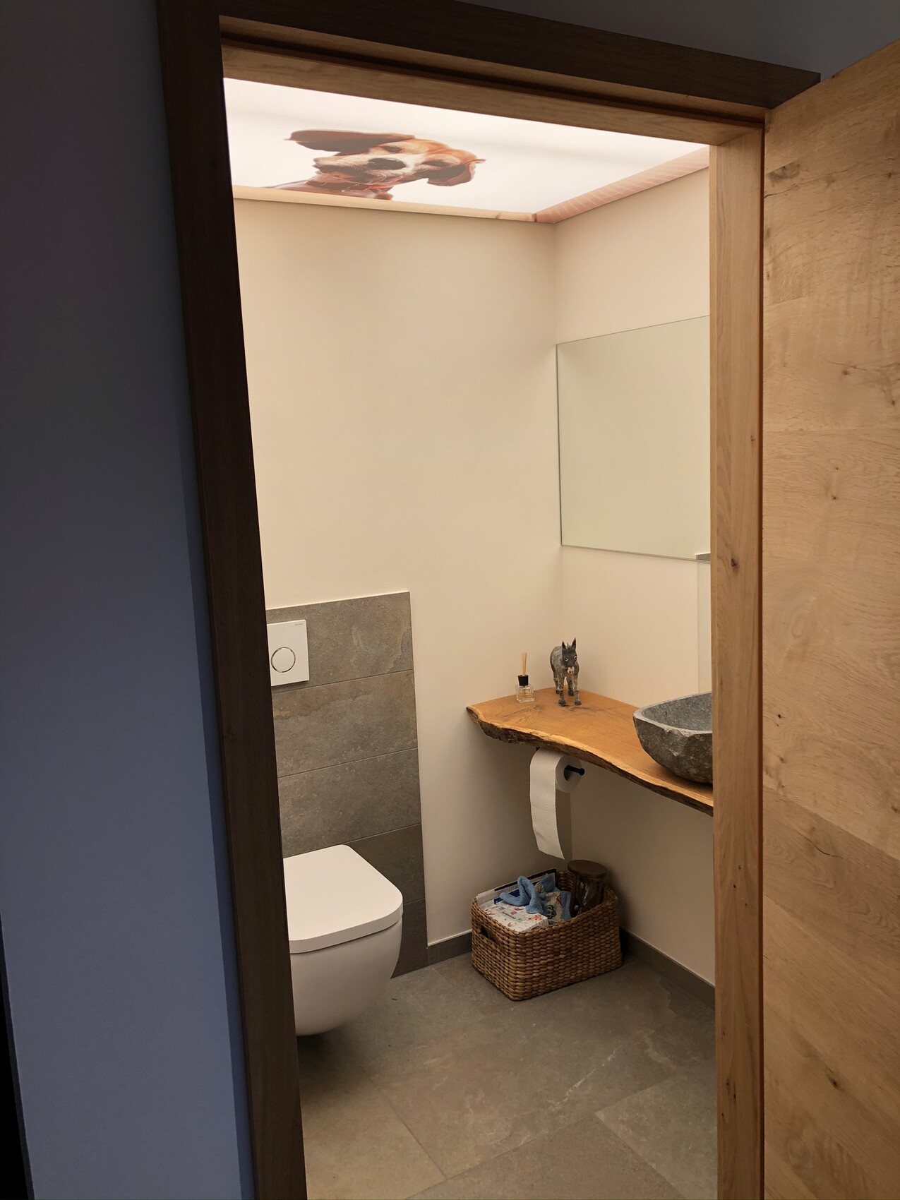 Bathroom with wall-hung toilet, wooden shelf, stone washbasin, cat figurine and towel basket.