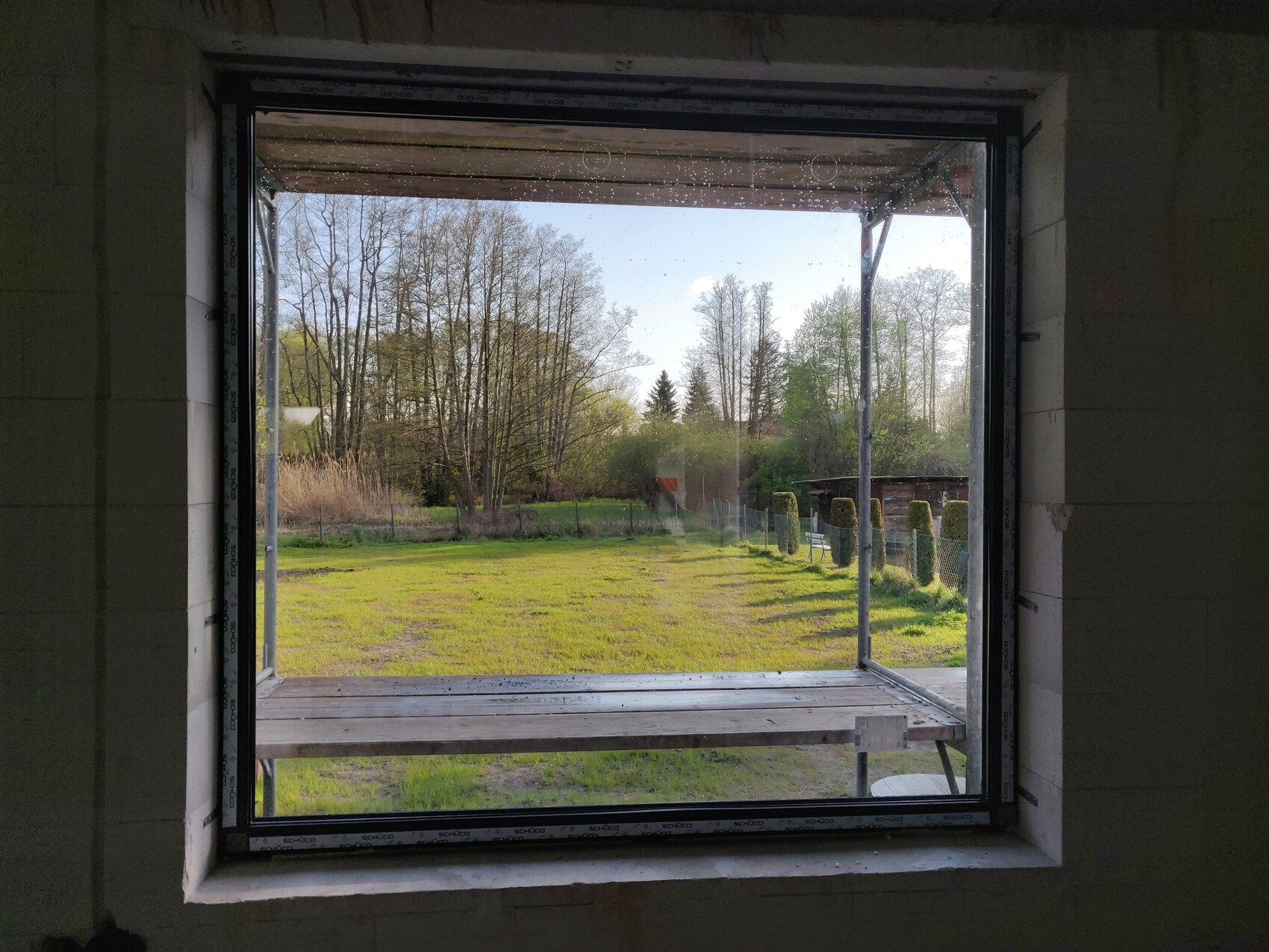 View from unfinished interior through open window onto green meadow, trees, fence, and wooden bench.