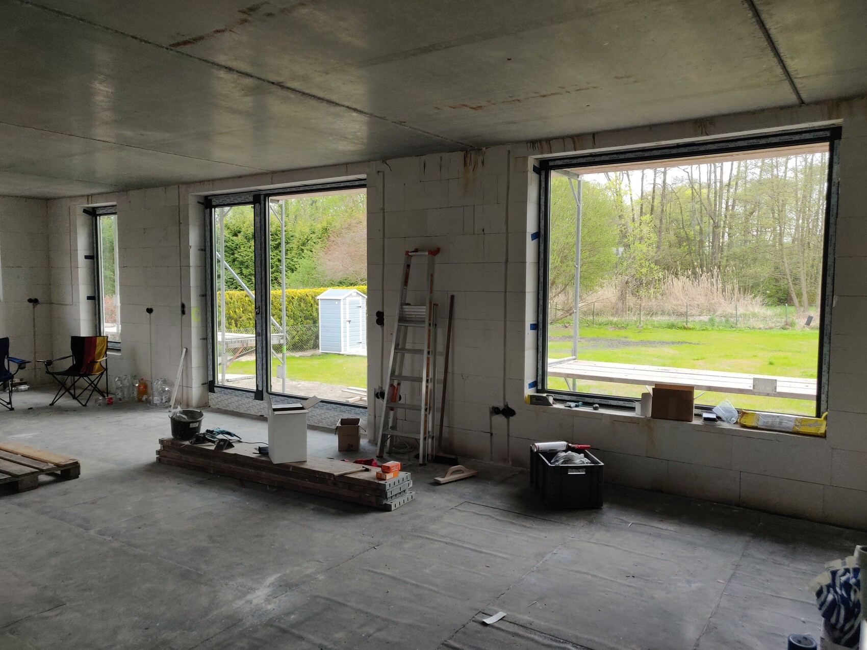 Unfinished interior of construction site with large windows, ladder, and stacks of wood.