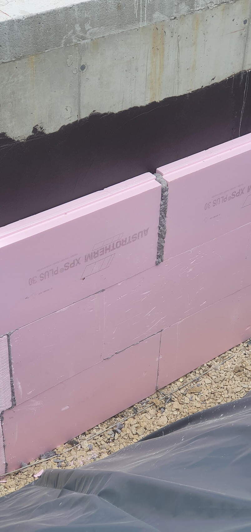 Construction wall: pink insulation panels (polystyrene) against concrete wall; gray joint mortar; gravel floor and construction foil.