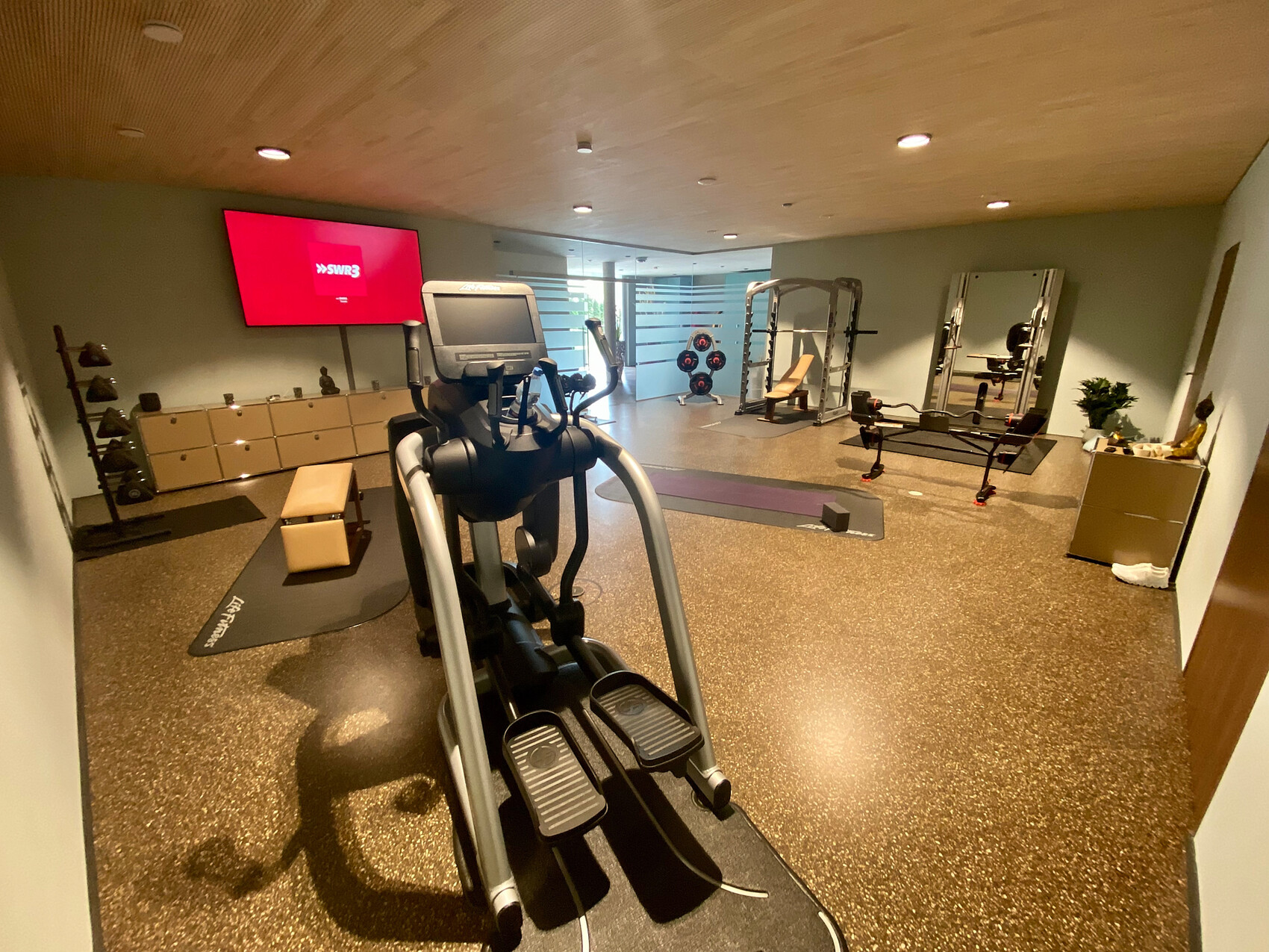 Elliptical trainer in gym room, weight bench, mirrored wall, weights, TV on the wall.