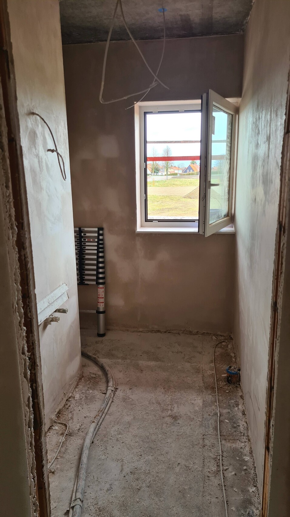 Unfinished construction space with open window front, pipes/cables, plaster; view outside to fields.