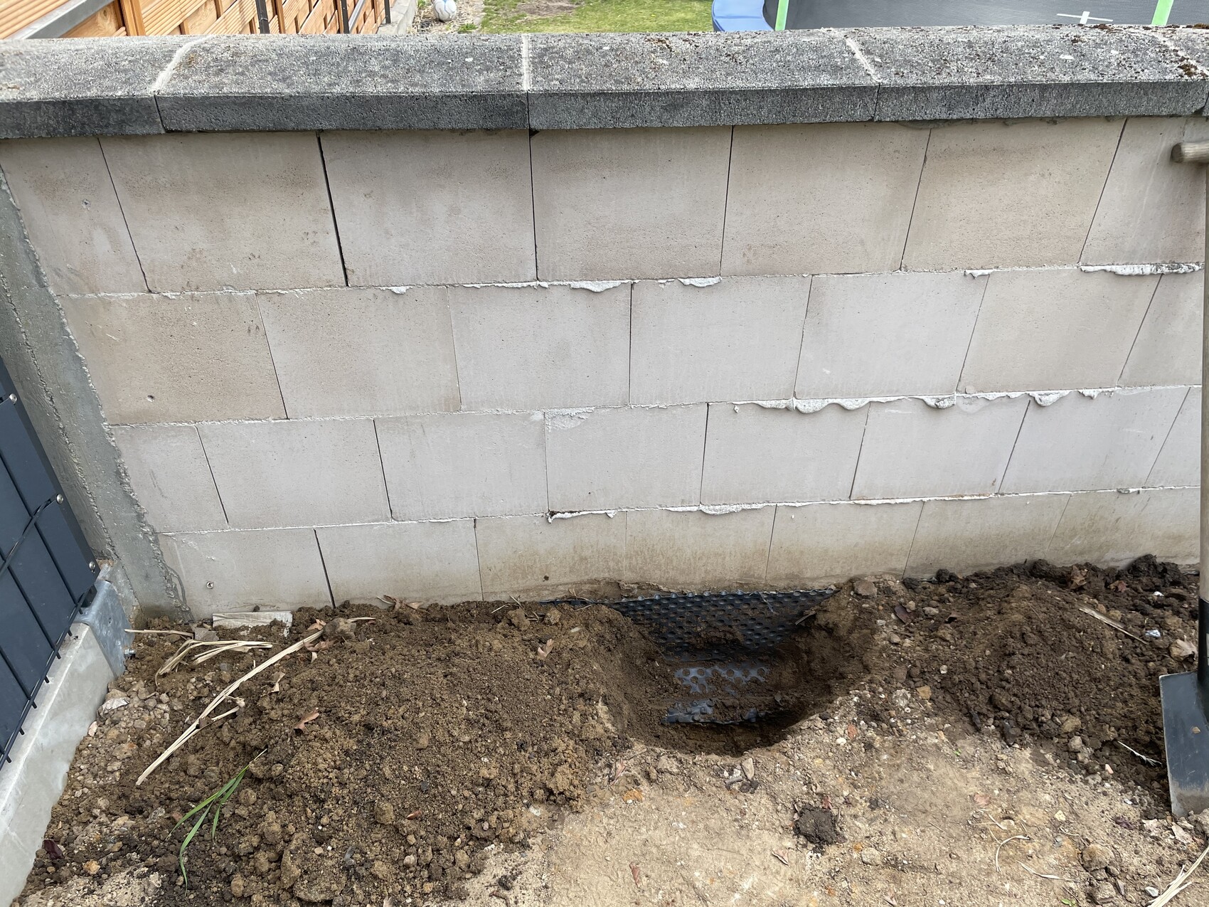 Gray concrete block wall, in front an open earth trench with black perforated sheet as drainage.