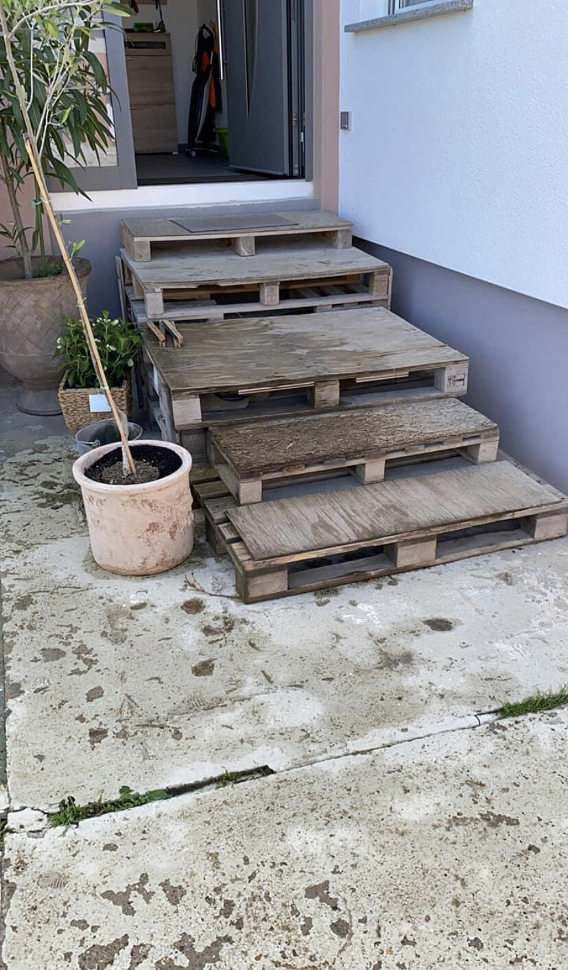 Mehrstufige Treppe aus Paletten vor offener Haustür; links Pflanze, nasser Betonboden.