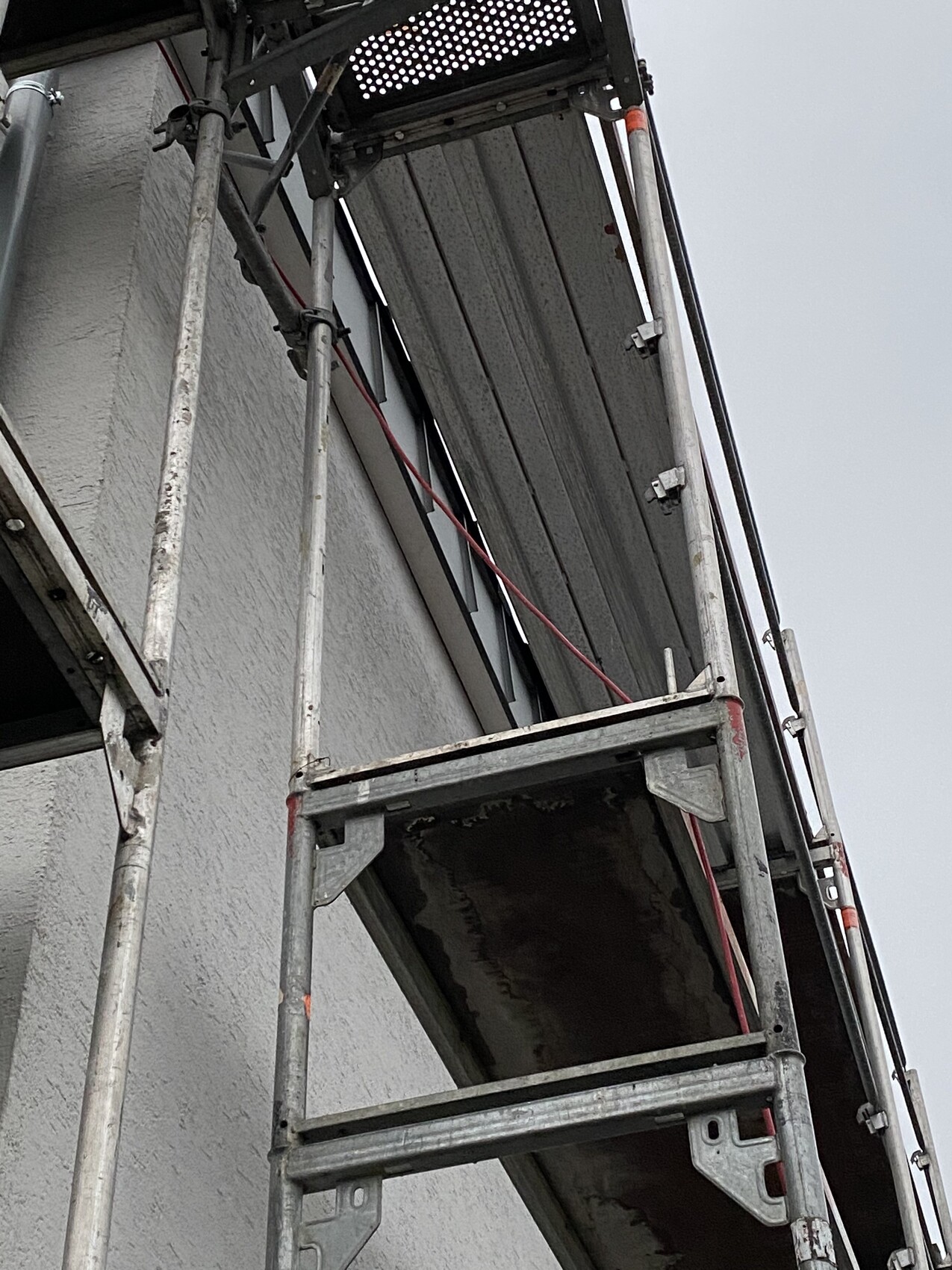 Metal scaffolding in front of a building wall with platform, railing, and pipes.