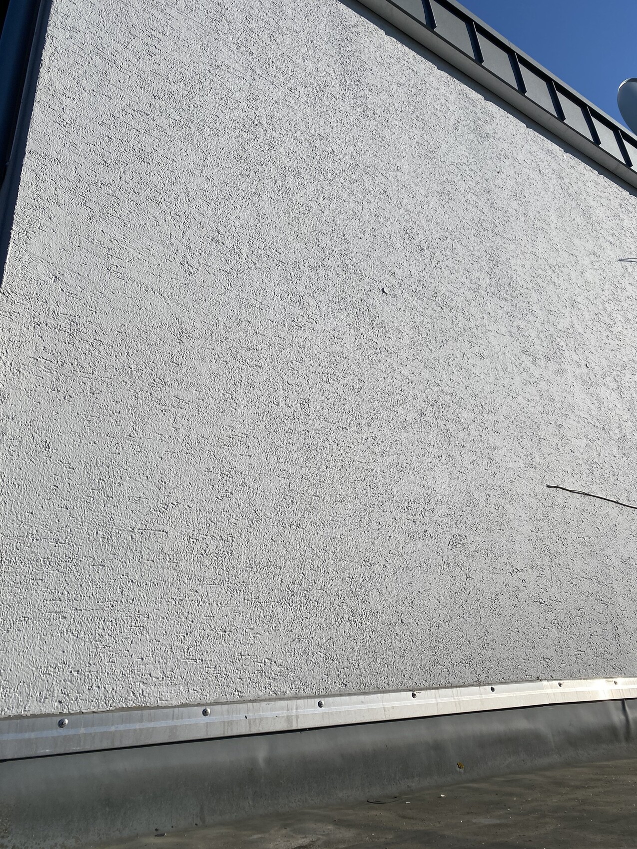 Weiße, grob strukturierte Außenwand eines Gebäudes mit Dachrand und blauem Himmel oben rechts.