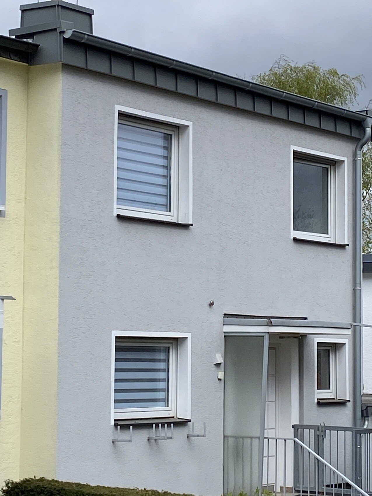 Zweistöckiges Wohnhaus mit grauer Putzfassade, weißen Fensterrahmen und Eingang mit Vordach