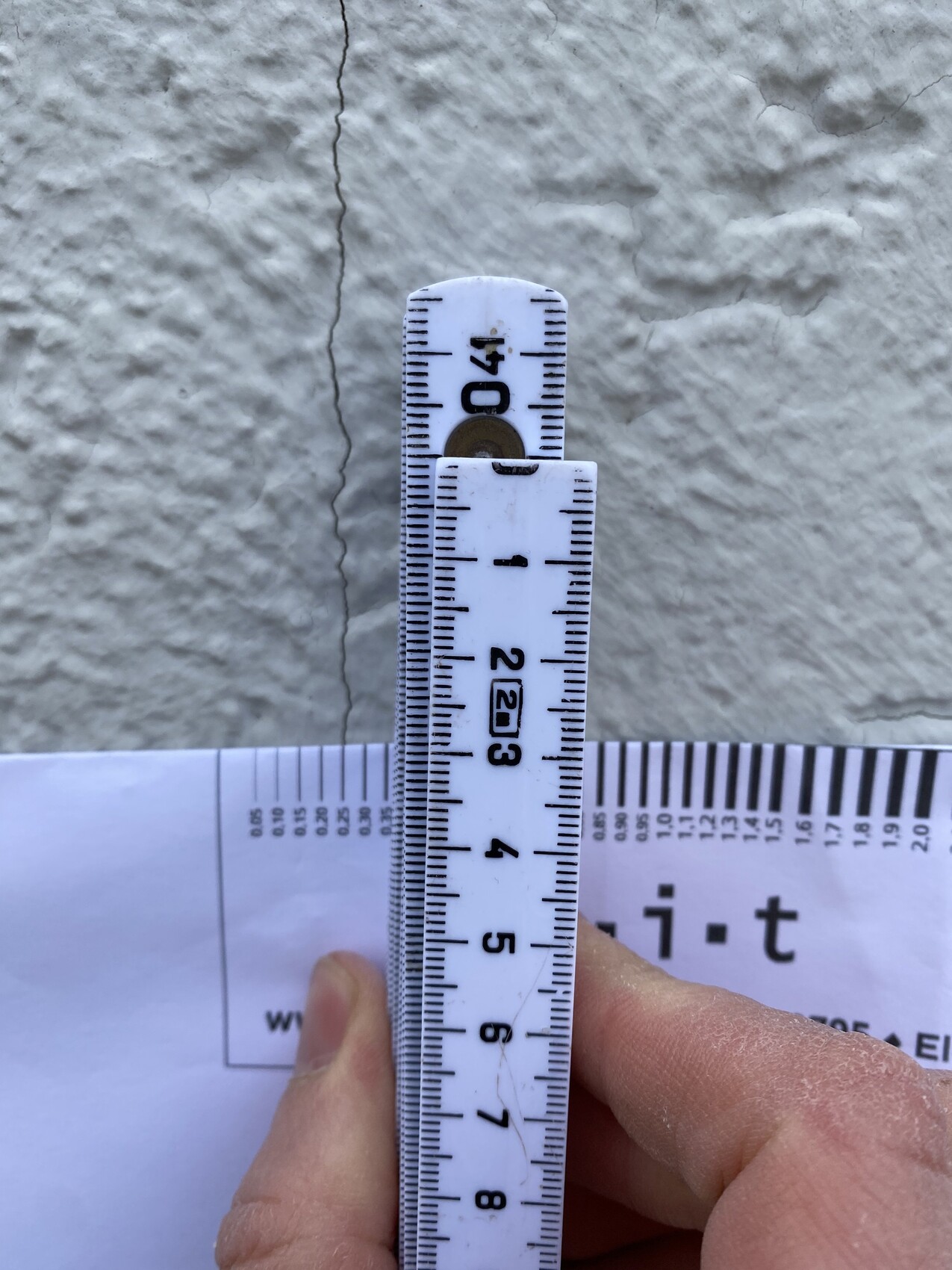 Nahaufnahme eines weißen Zollstocks vor rauer Wand mit vertikalem Riss; Hand hält das Lineal.