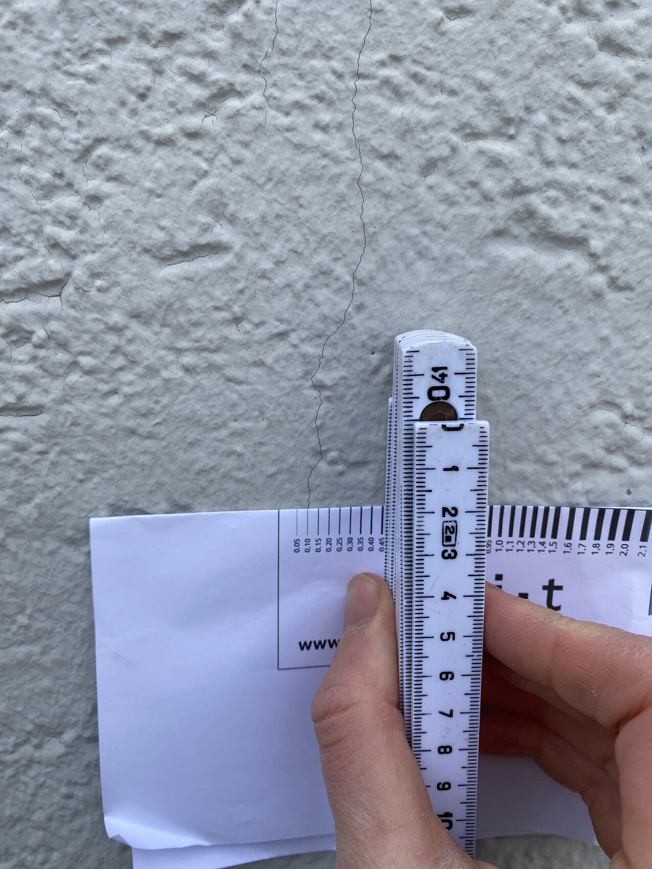 Eine Hand misst mit einem Faltruler die Breite eines Risses in einer rauen weißen Wand.