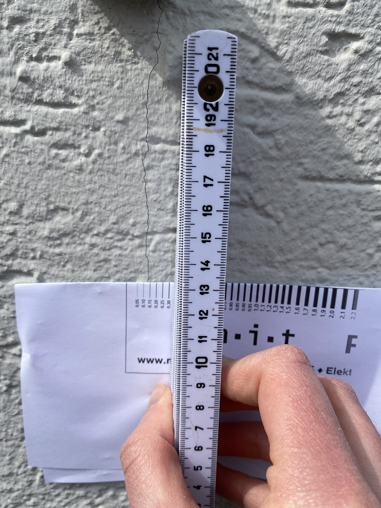 Maßband senkrecht an rauer Steinwand, Hand hält Band, darunter Papier mit URL.