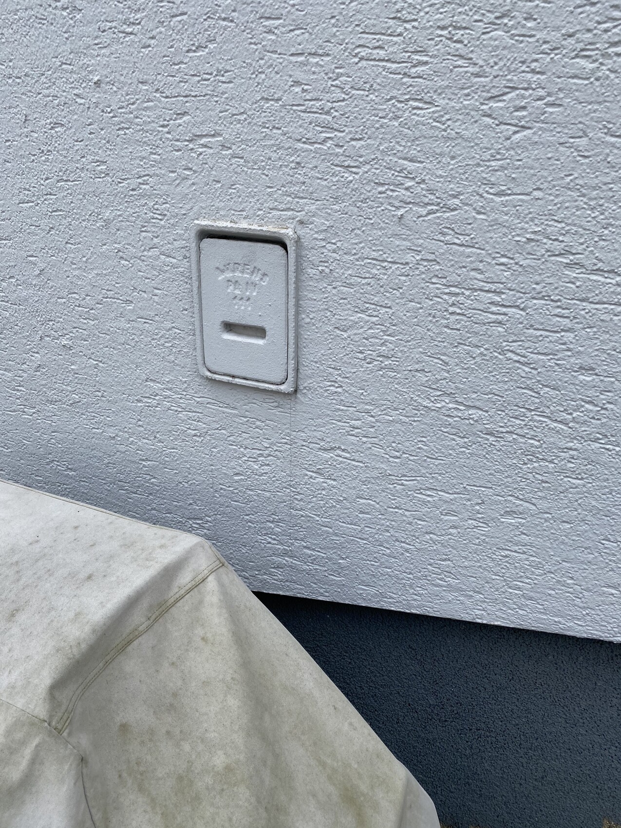 Quadratische Wandklappe mit Schlitz an rauer hellgrauer Außenwand; davor eine beige Stoffabdeckung.