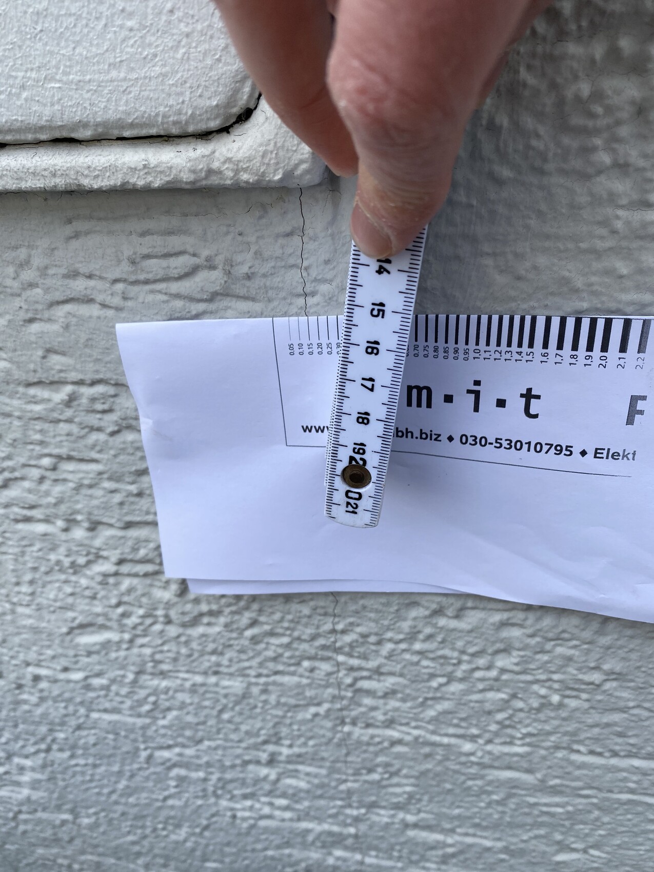 Eine Hand hält ein Maßband gegen eine Wand, um einen Riss zu messen.