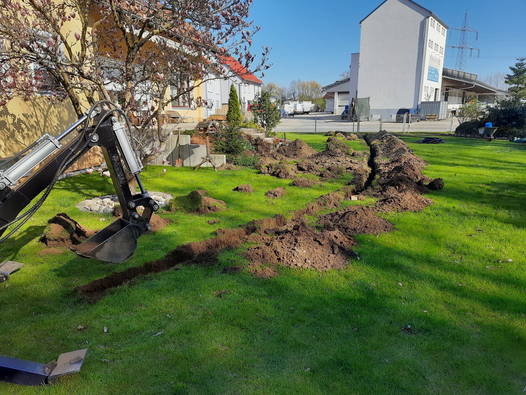 Excavator digging a long trench in the garden grass; piles of soil line the excavation.