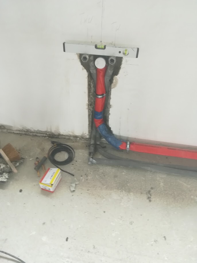 Wall opening with red and blue pipe system, spirit level above, tools on the floor.