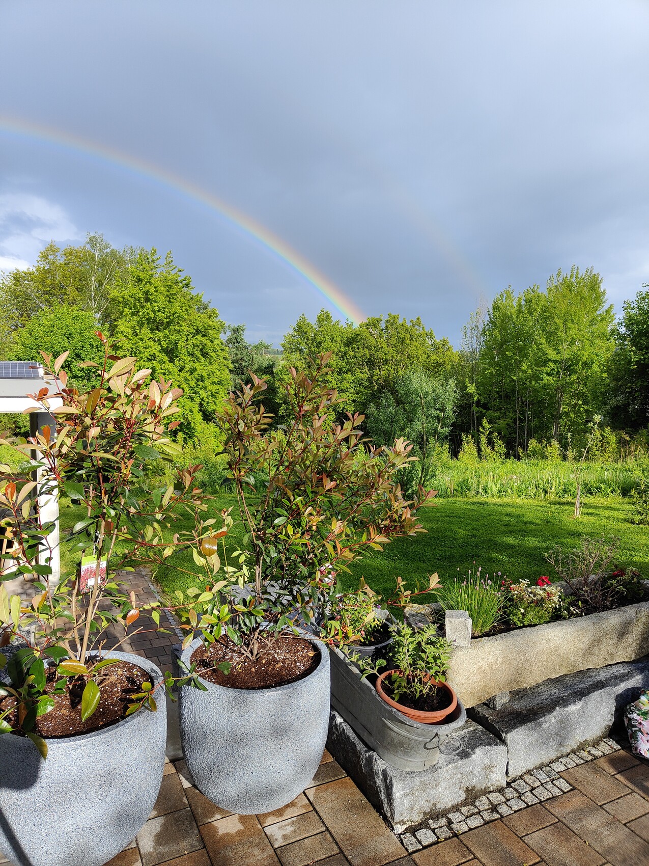 Doppelter Regenbogen über Garten; vorn Topfpflanzen auf Terrasse.