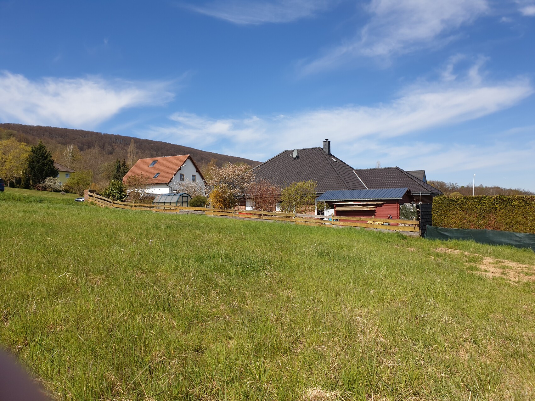 Green field in front of residential houses with wooden fence, trees and hills under blue sky.