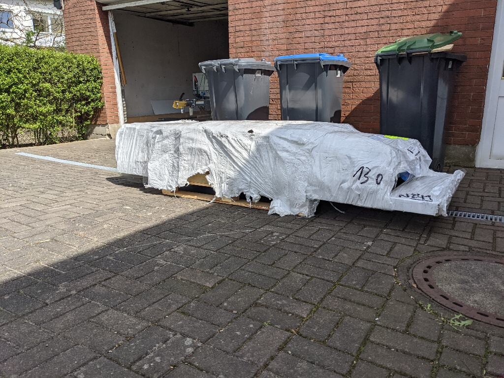 Long pallet covered with a tarpaulin in the yard in front of the garage; trash bins behind.