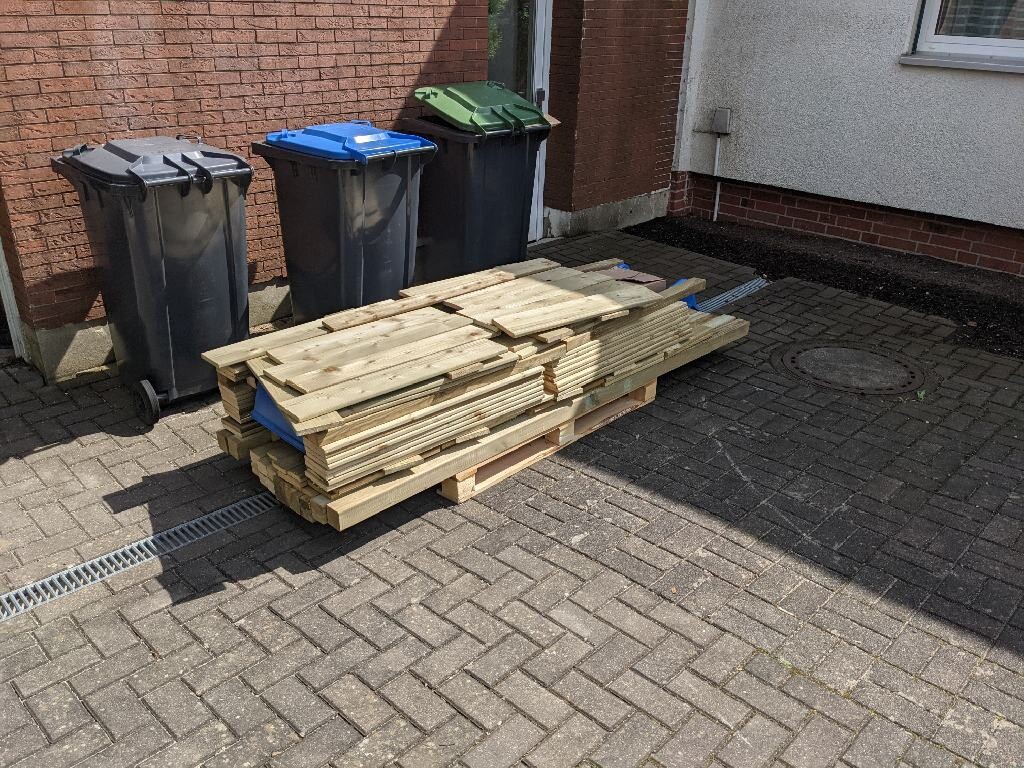 Wooden battens stacked on a pallet next to trash bins in front of the house facade.