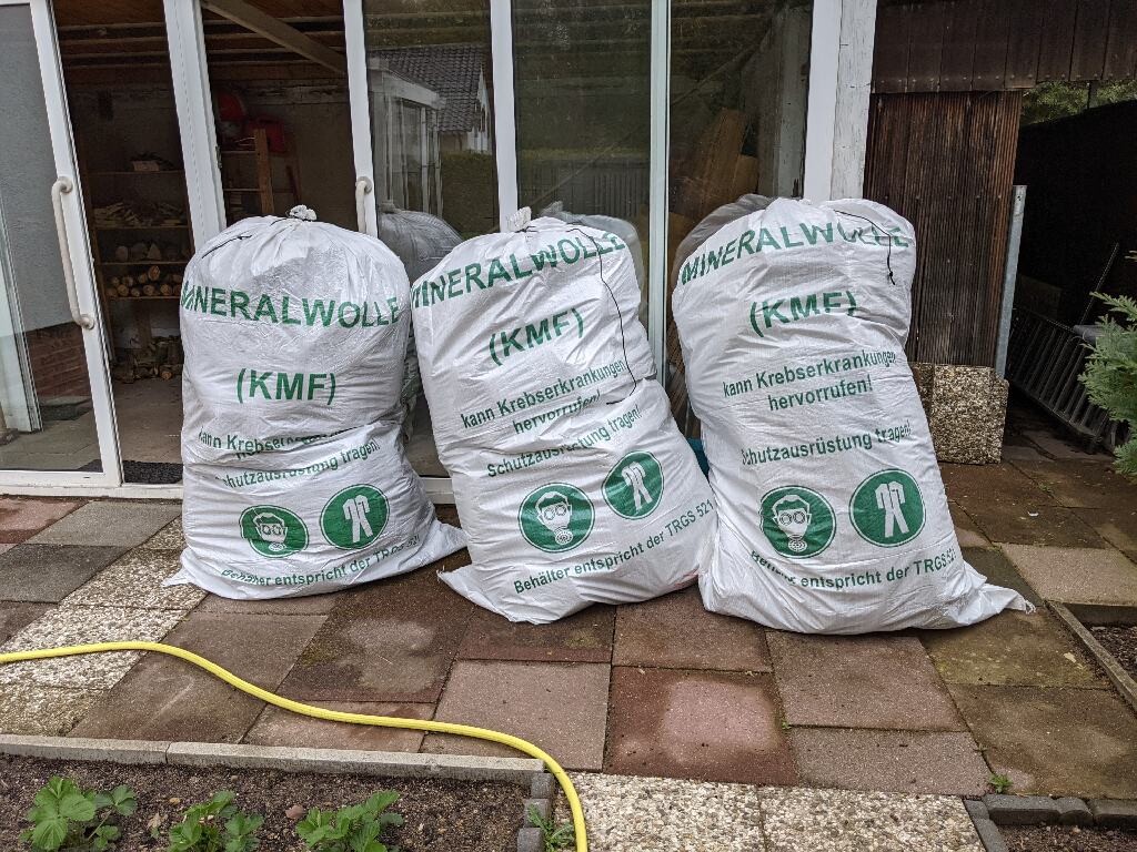 Three white bags of mineral wool insulation (MWI) standing in front of a glass door, yellow hose lying on the terrace.