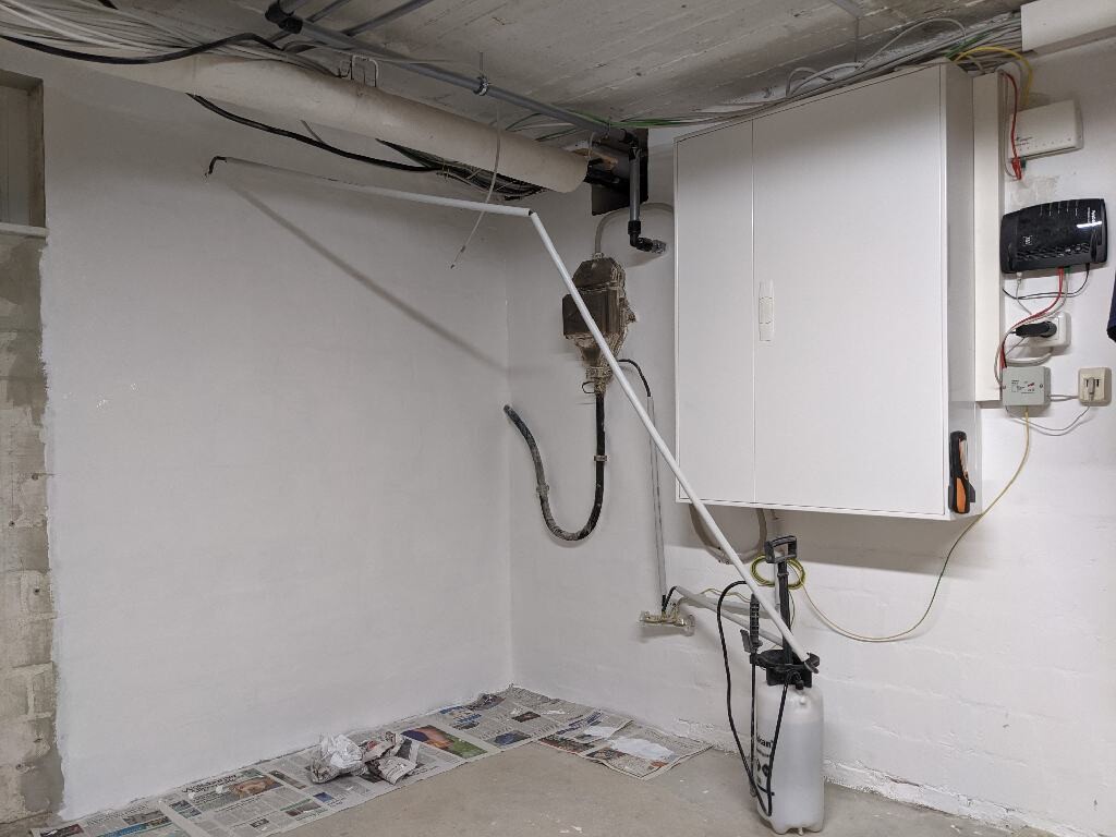 Basement room with white cabinet, pipes, cables, pump and a newspaper on the floor.
