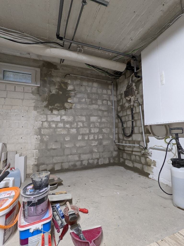Basement room with unfinished brick wall, heating system, pipes and tools on the floor.