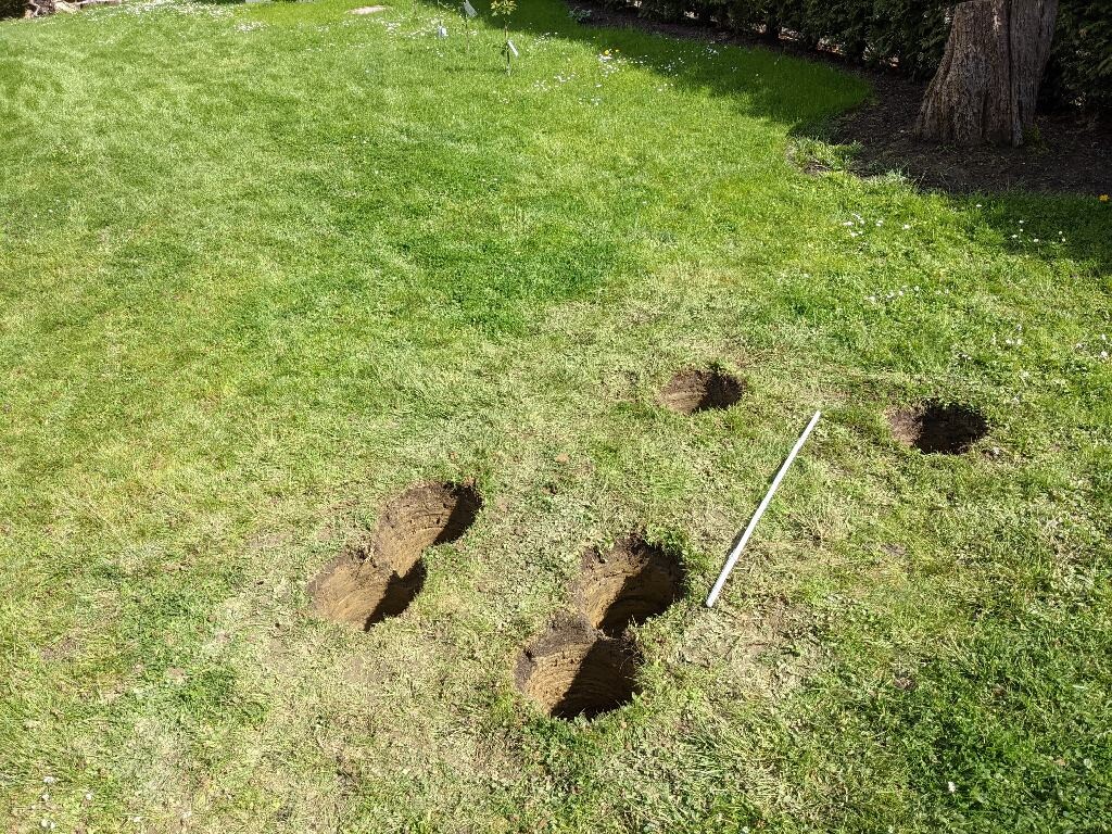 Lawn area with several open holes in the ground; a white measuring rod is lying on the grass.