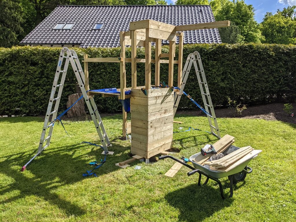 Wooden play structure in the garden under construction, ladders on the side, wheelbarrow full of wooden parts.