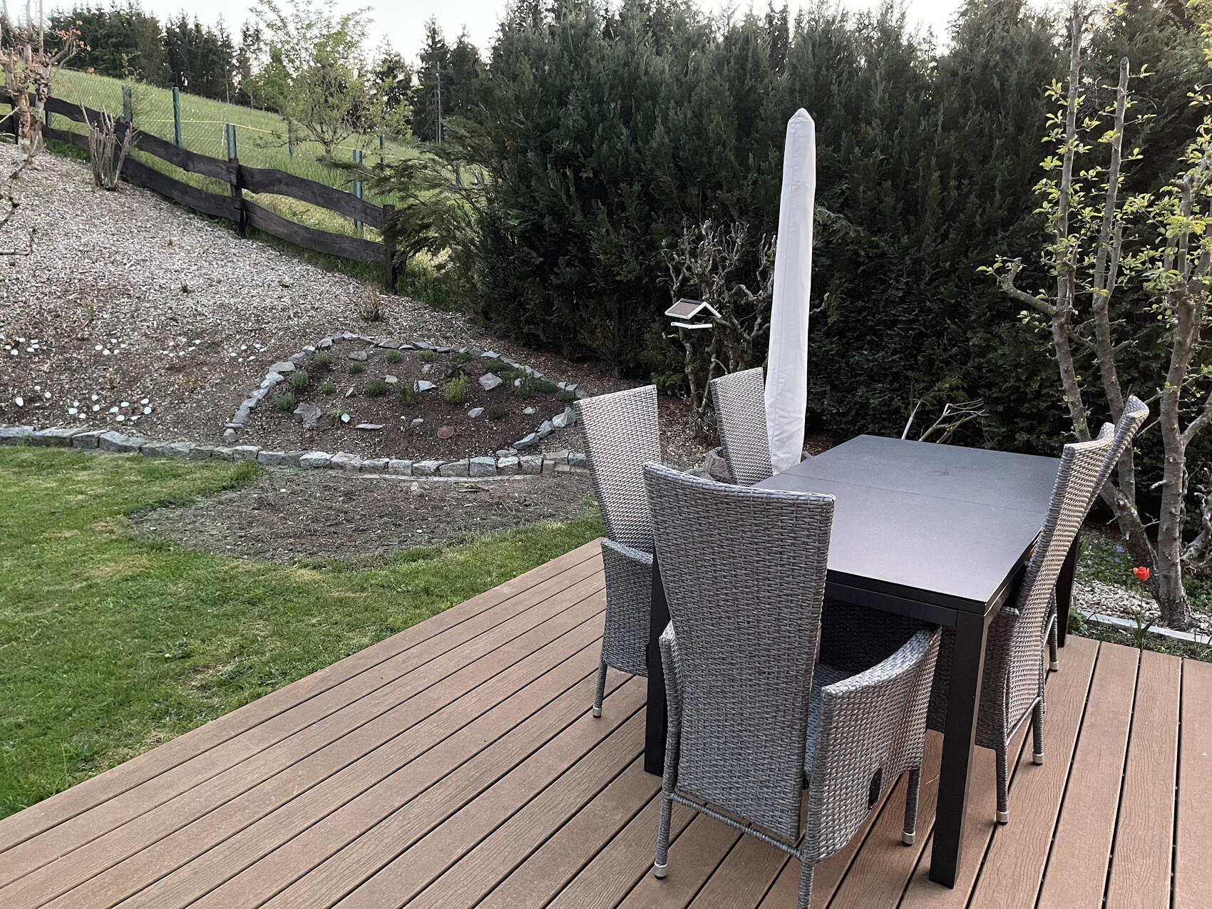 Wooden deck with gray garden table and wicker chairs, closed white parasol, hedge behind.