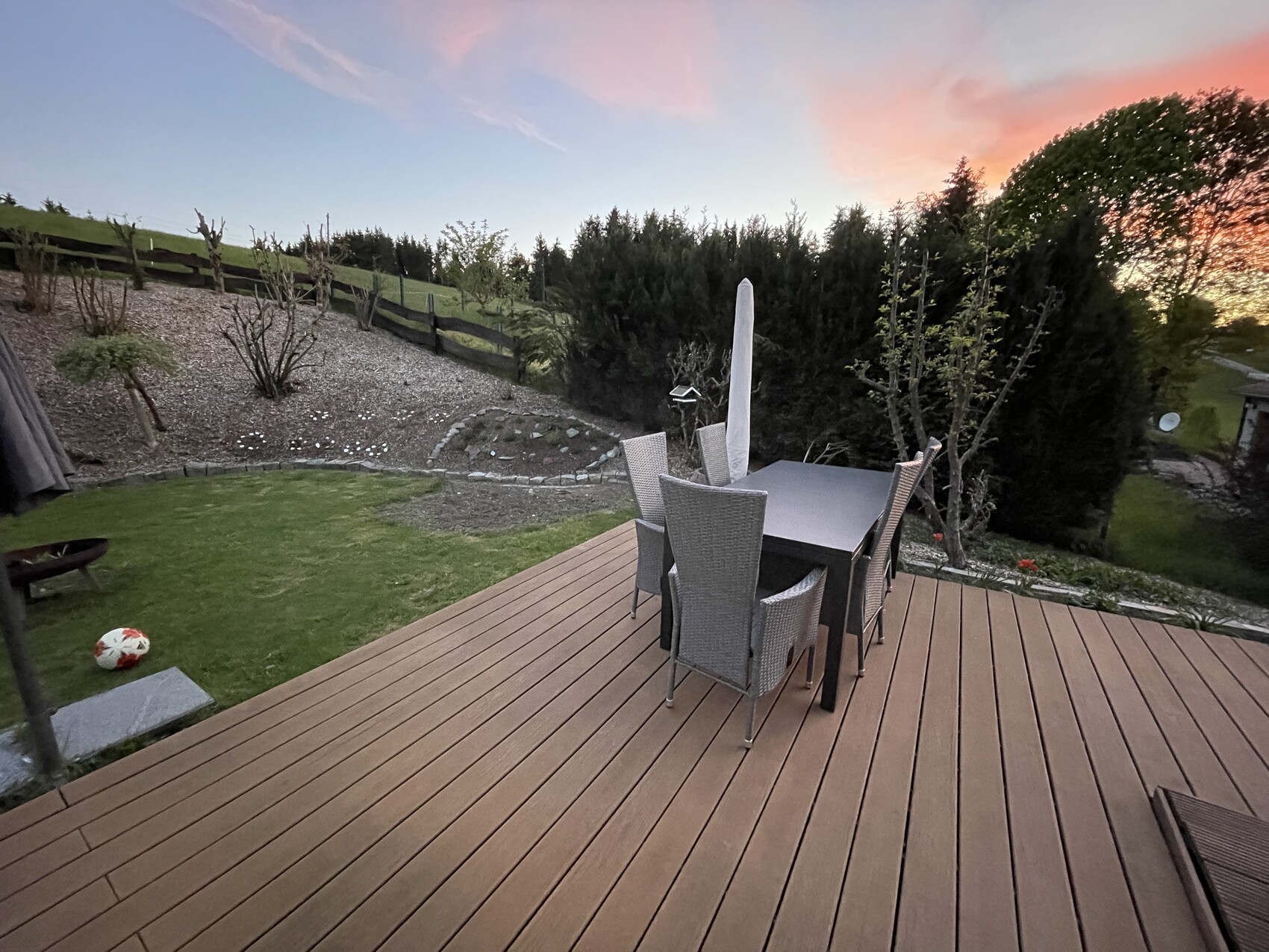 Wooden deck with dining table and chairs, closed parasol, garden in evening light.