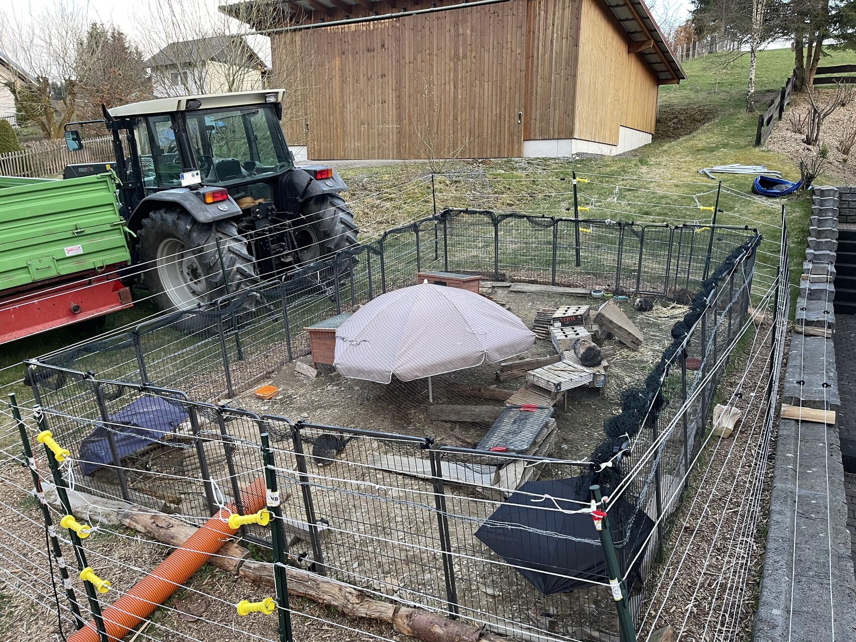 Garden enclosure with metal fence, tractor on the left, round umbrella and firewood in the yard.