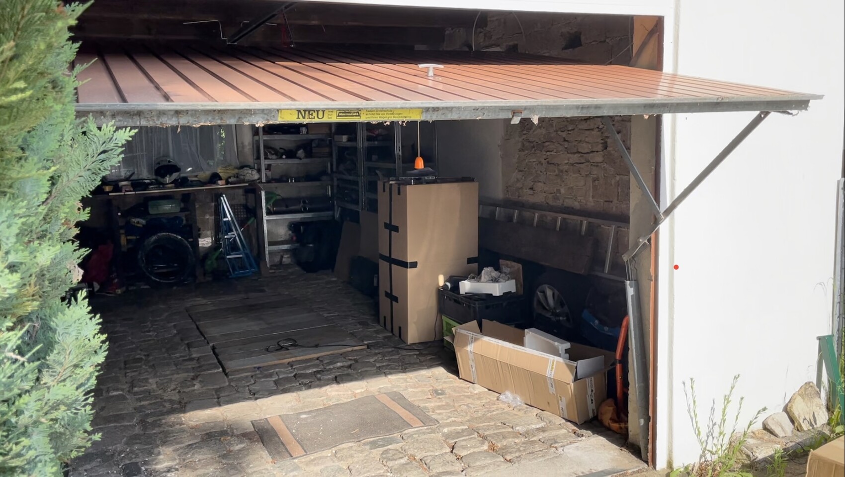 Open garage with workbench, shelves, boxes, toolboxes, and ladder; stone floor.