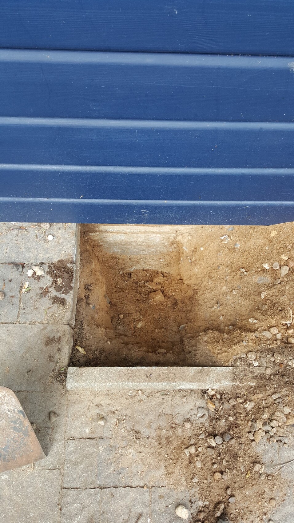 Offene Fundamentgrube neben blauer Fassadenwand auf Baustelle mit Erde und Kies