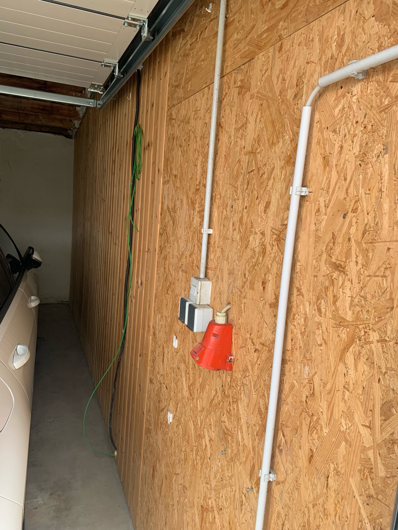 Garage mit OSB-Wand, roter Elektroanschluss, weiße Leitungen, grüner Schlauch, Auto am Rand.