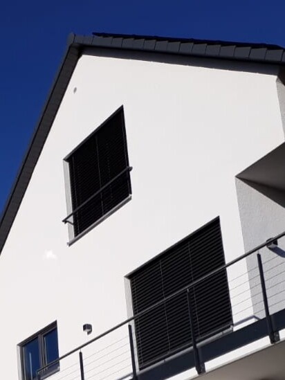 White modern house facade with black shutters, balcony with wire cable railing, blue sky.