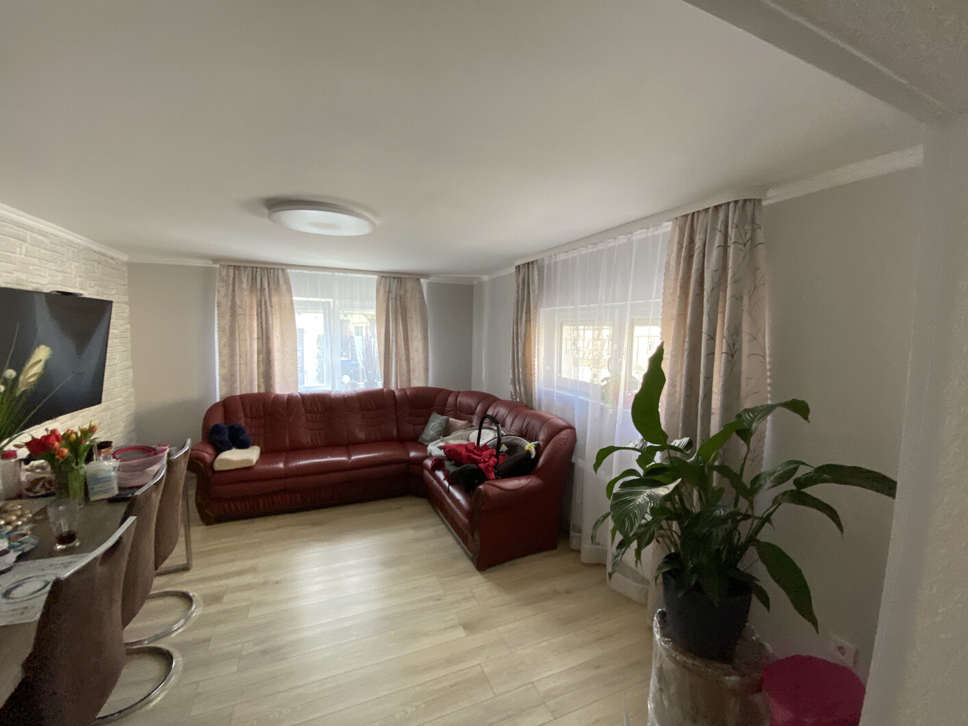 Living room with red corner leather sofa, light curtains, plant on the right, dining table on the left.