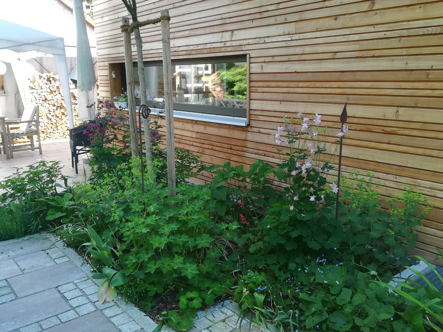 Garden bed next to wooden house with blooming flowers, stone path, patio, and woodpile in the background.