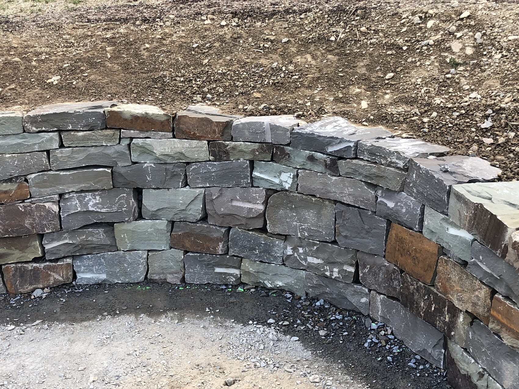 Curved dry stone wall made of irregular natural stones in front of an earth embankment.