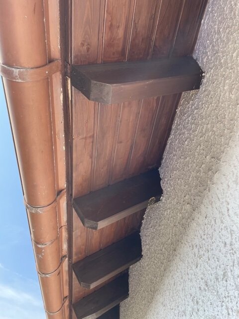 Wooden wall with three protruding boards as steps; brown downpipe on the left, plastered wall on the right.