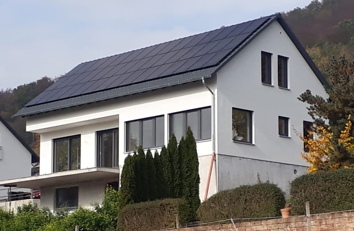 Modernes zweistöckiges Haus mit Solpanelen auf dem Dach, große Fensterfront und Hecke vor dem Haus.