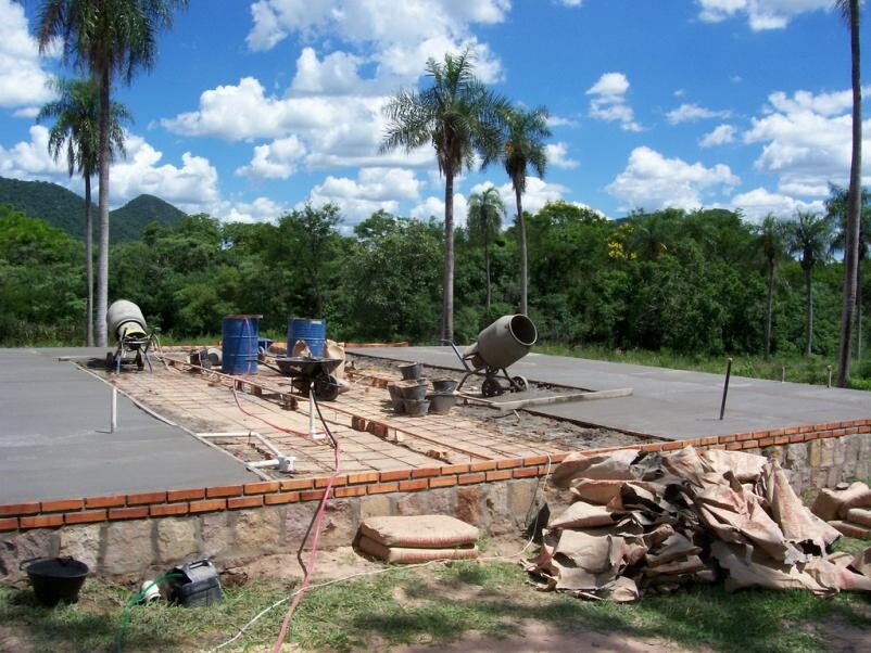 Construction site with foundation slab, concrete mixer and cement bags; palm trees and forest in the background.
