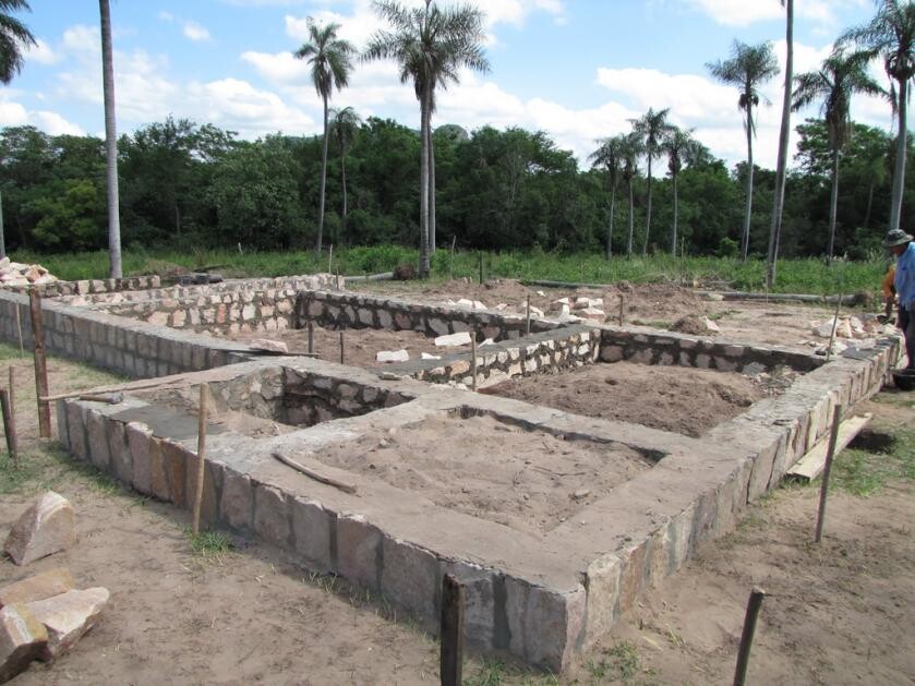 Foundation made of rubble stone walls on construction site; workers on the right, palm trees and forest in the background.