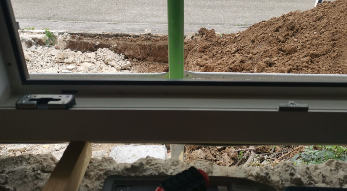 View through window onto a construction site: soil, rubble, green pole, tools in foreground.