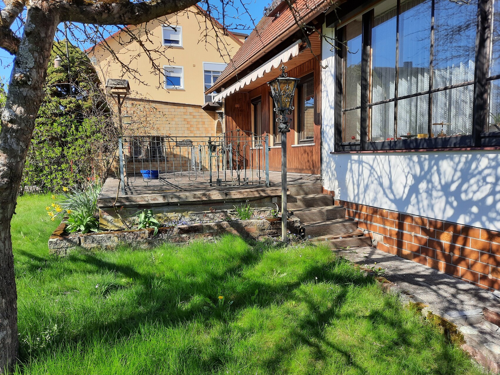 Garden with wooden deck, railing and stairs leading to the house; lantern, large windows, tree in the foreground.